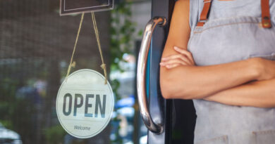 Small Business - Woman standing beside "Open" sign with arms crossed.