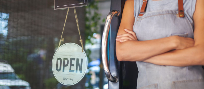 Small Business - Woman standing beside "Open" sign with arms crossed.