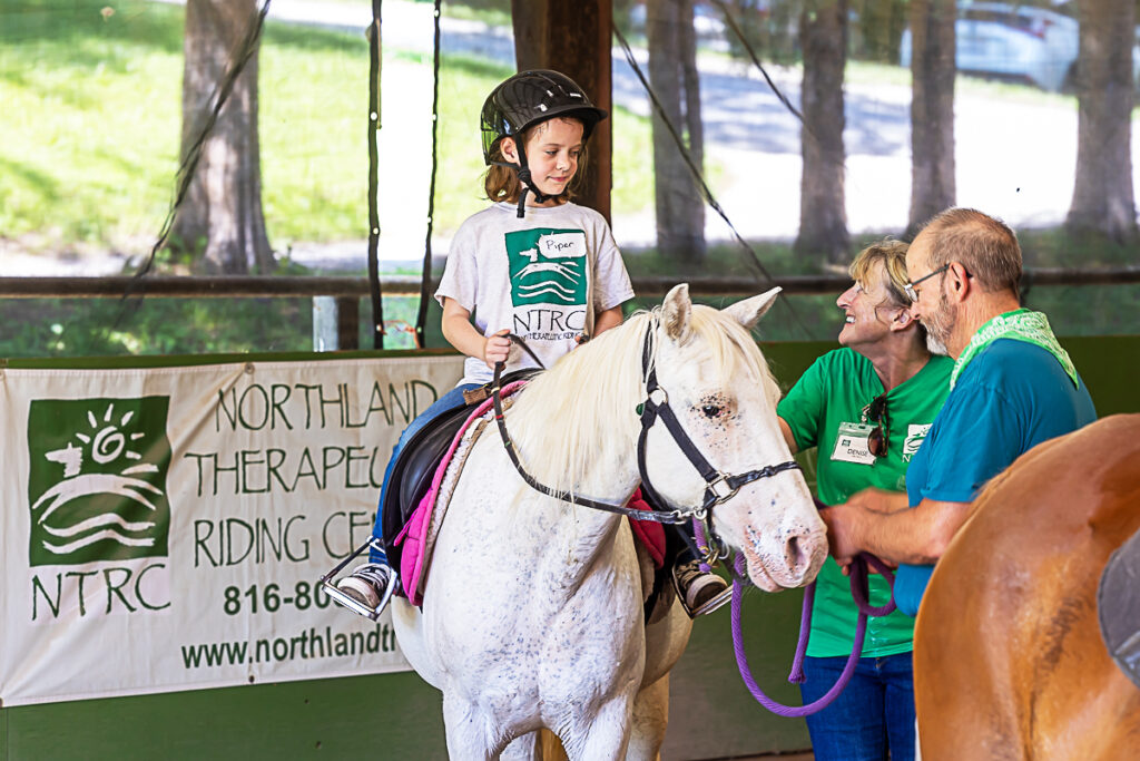 Photograph of a child riding a white horse with two trainers leading the horse at the Northland Therapeutic Riding Center in Kearney, Missouri.