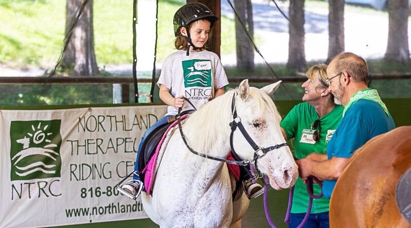 Photograph of a child riding a white horse with two trainers leading the horse at the Northland Therapeutic Riding Center in Kearney, Missouri.