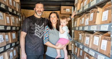 Scott and Jess stand in a delivery trailer holding their young daughter, surrounded by shelves of boxes.