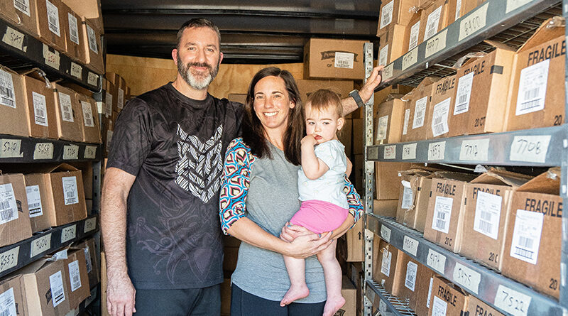 Scott and Jess stand in a delivery trailer holding their young daughter, surrounded by shelves of boxes.