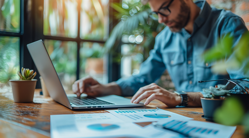 Man working on business paperwork and laptop at desk surrounded with plants.