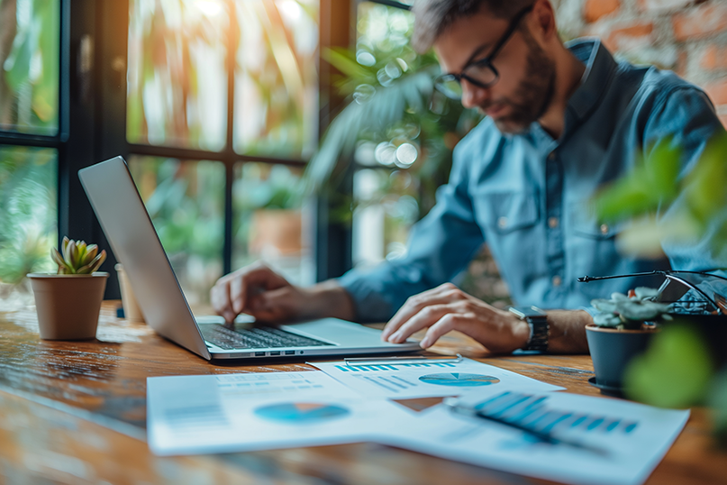 Man working on business paperwork and laptop at desk surrounded with plants.
